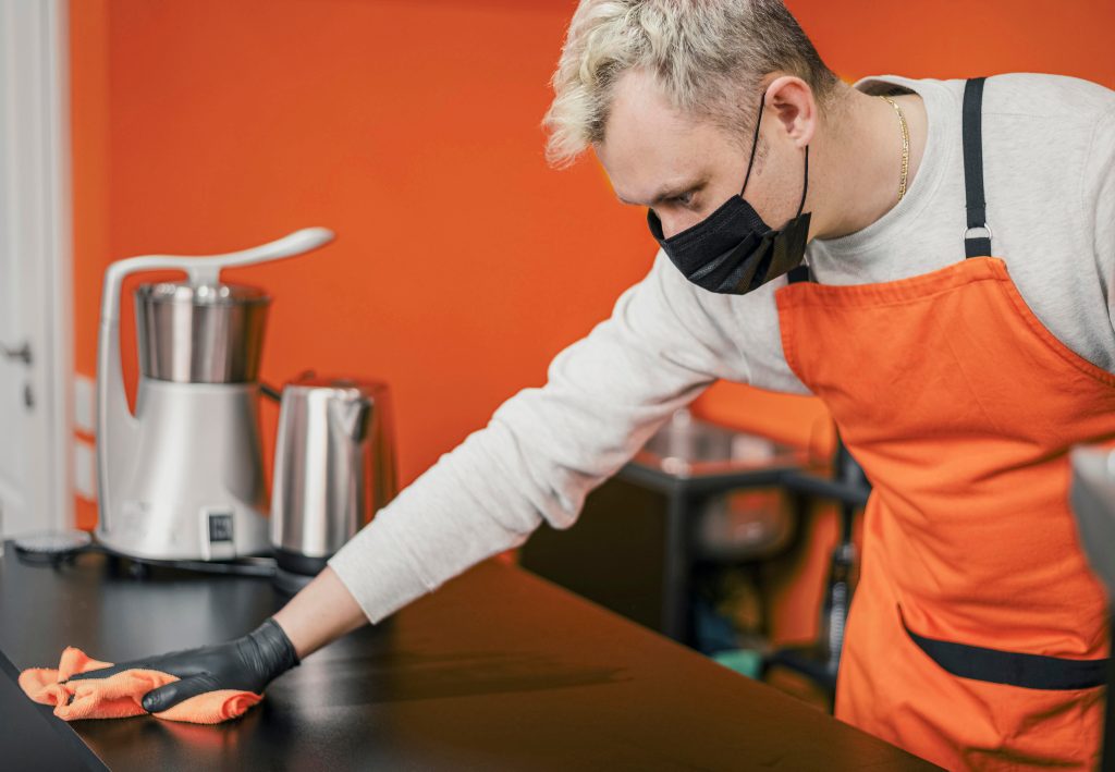 A restaurant employee wearing an apron and face mask disinfects a countertop as part of routine cleaning and sanitation.